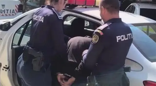 Doi polițiști în uniformă întunecată stau lângă o mașină albă, unul dintre ei având o pistol în toc la brâu.