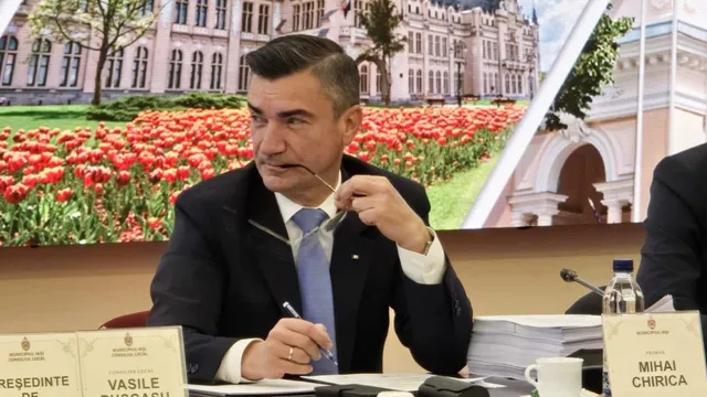 Un bărbat într-un costum închis și cravată deschisă stă la o masă cu documente, ținând ochelari și un pix.
