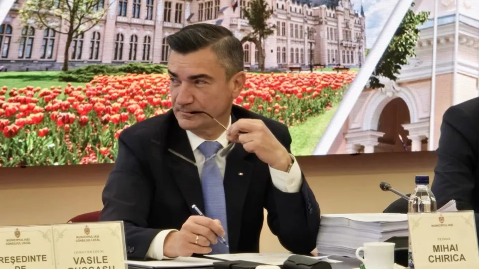 Un bărbat într-un costum închis și cravată deschisă stă la o masă cu documente, ținând ochelari și un pix.