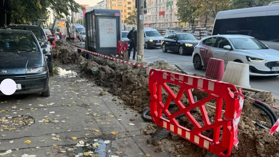 Un șantier cu pământ și bariere de protecție roșii se află pe un trotuar lângă mașini parcate și un autobuz.