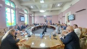 Oamenii stau așezați la o masă mare, ovală, într-o sală cu pereți roz și ferestre înalte cu vitralii colorate.
