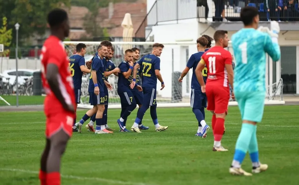 Un grup de fotbaliști în echipament albastru se bucură pe un teren de fotbal, în timp ce jucători în echipament roșu și turcoaz stau în apropiere.