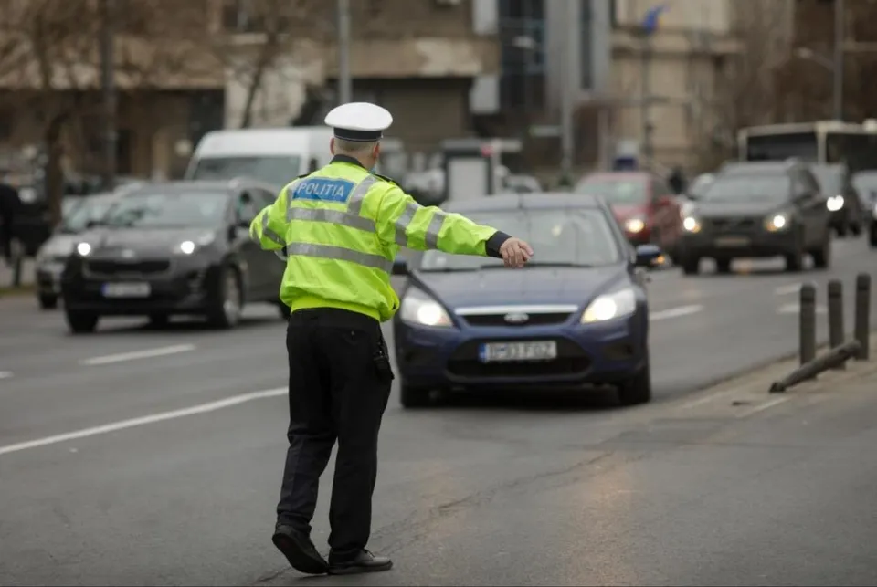 Un polițist cu jachetă reflectorizantă stă pe carosabil și întinde brațul spre mașini oprite în trafic urban.