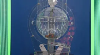 O sferă transparentă de loto conține bile colorate și este montată pe un suport de plastic transparent în fața unui fundal albastru.