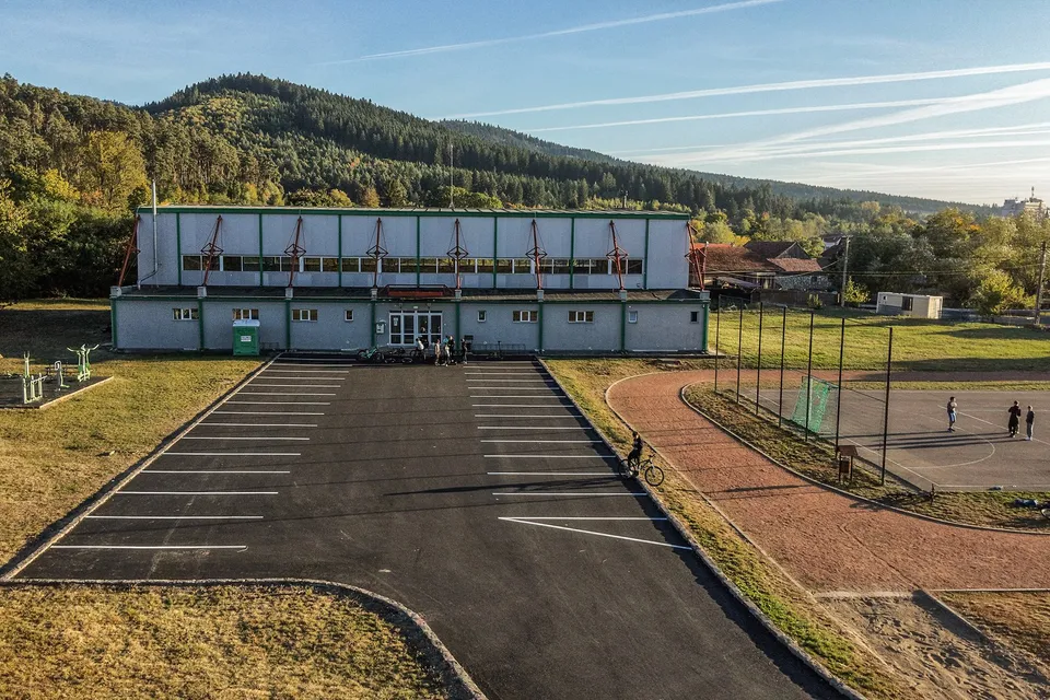 O clădire gri cu ferestre lungi se află lângă un teren de sport și un teren de asfalt cu linii albe marcate pentru parcare.
