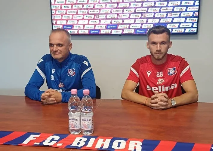 Un bărbat în echipament sportiv albastru și un bărbat în echipament sportiv roșu stau la o masă cu două sticle de apă în față.