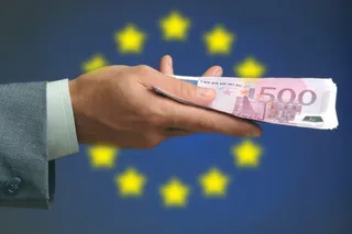 O mână într-un sacou gri ține o bancnotă roz de cinci sute de euro în fața unui fundal albastru cu stele galbene.