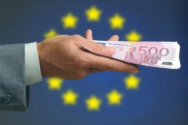 O mână într-un sacou gri ține o bancnotă roz de cinci sute de euro în fața unui fundal albastru cu stele galbene.