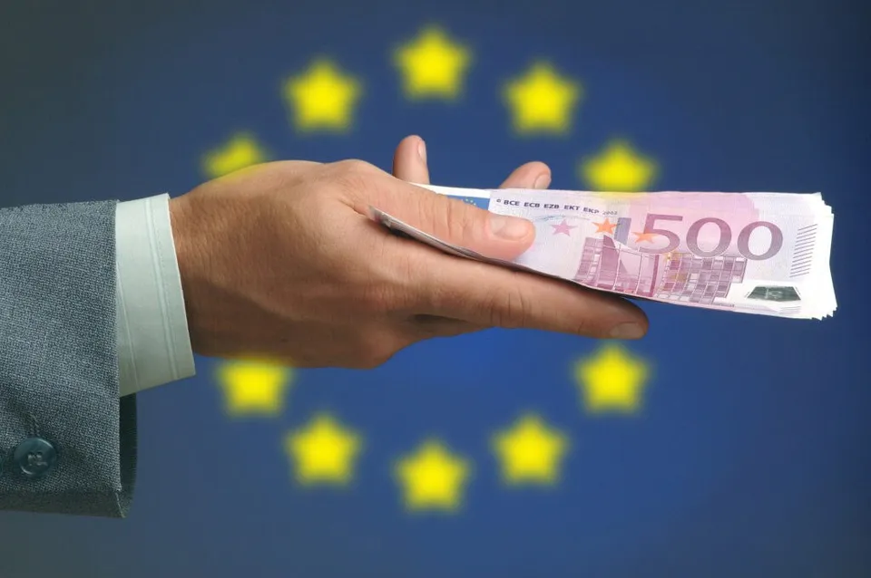 O mână într-un sacou gri ține o bancnotă roz de cinci sute de euro în fața unui fundal albastru cu stele galbene.