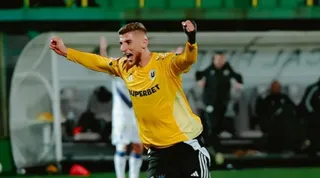 Un bărbat cu tricou galben și pantaloni negri stă cu brațele ridicate și mâinile strânse în pumni pe un teren de fotbal.