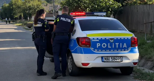 Doi polițiști stau lângă portbagajul deschis al unei mașini de serviciu albastră și albă cu inscripția politia și girofaruri aprinse.