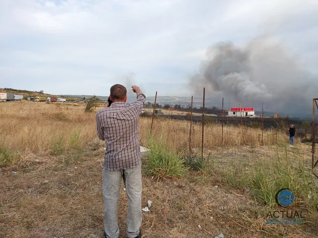 Un bărbat cu cămașă în carouri și pantaloni deschiși stă cu spatele, arătând spre un nor de fum de pe un câmp cu iarbă uscată.