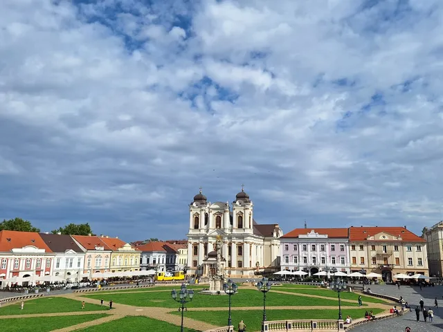 O clădire barocă cu două turnuri se ridică deasupra unei piețe cu o zonă verde centrală și clădiri cu acoperișuri roșii.