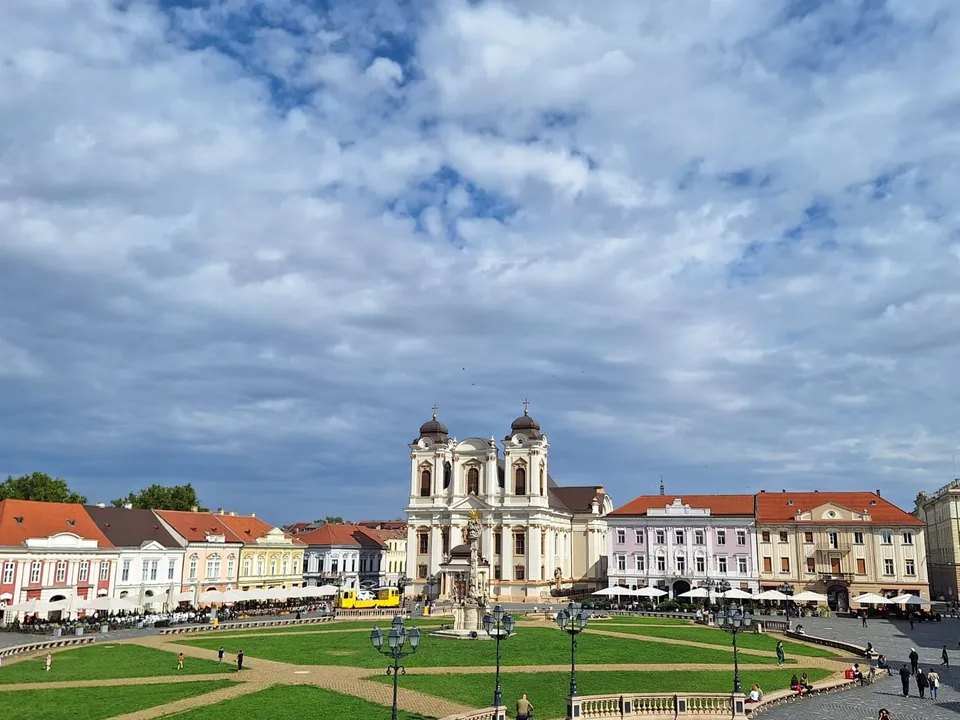 O clădire barocă cu două turnuri se ridică deasupra unei piețe cu o zonă verde centrală și clădiri cu acoperișuri roșii.