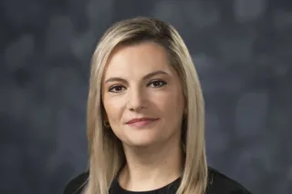 O femeie cu părul blond și bluză neagră privește spre cameră pe un fundal gri neclar.