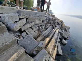Blocuri de beton și materiale inerte sunt depozitate pe malul unui corp de apă lângă câțiva oameni care stau în picioare.