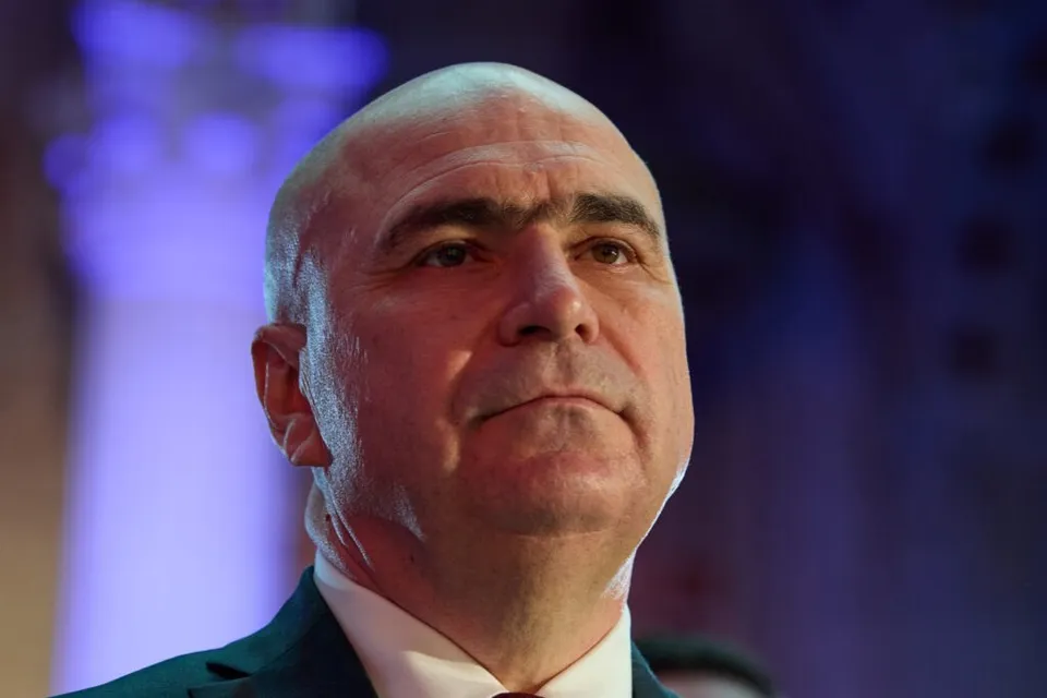 Un bărbat cu capul chel și pielea deschisă poartă o cămașă albă și un sacou verde în fața unui fundal albastru.