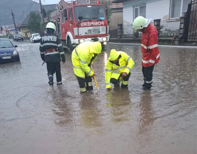 Patru persoane purtând haine de protecție și căști stau pe o stradă inundată lângă un camion de pompieri roșu.