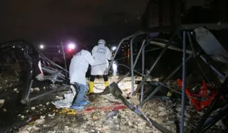 Două persoane îmbrăcate în haine albe stau lângă structuri metalice deformați și resturi pe un teren noaptea.