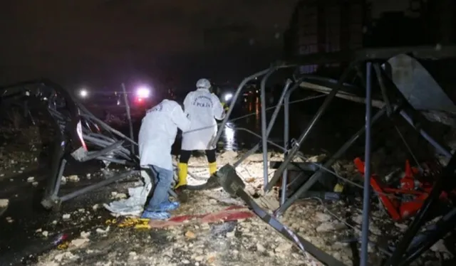 Două persoane îmbrăcate în haine albe stau lângă structuri metalice deformați și resturi pe un teren noaptea.