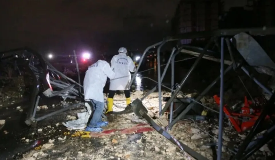 Două persoane îmbrăcate în haine albe stau lângă structuri metalice deformați și resturi pe un teren noaptea.