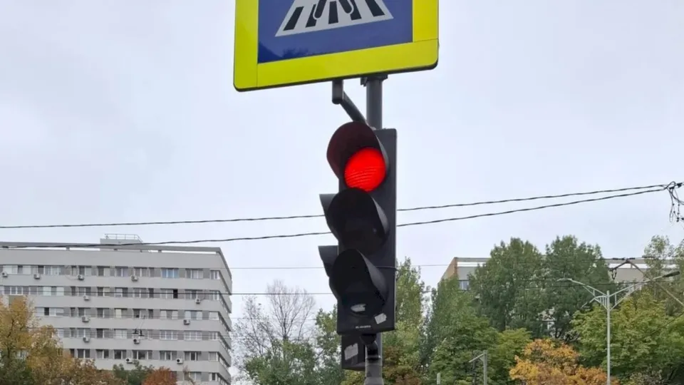 Un semafor cu lumina roasă aprinsă este montat pe un stâlp sub un panou de semnalizare rutieră galben cu pictograma trecerii de pietoni.