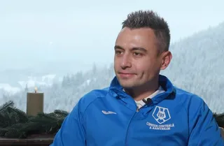 Un bărbat cu părul scurt și scurt, de culoare închisă, poartă un hanorac albastru cu un logo alb pe piept, stând în fața unui peisaj montan înzăpezit.