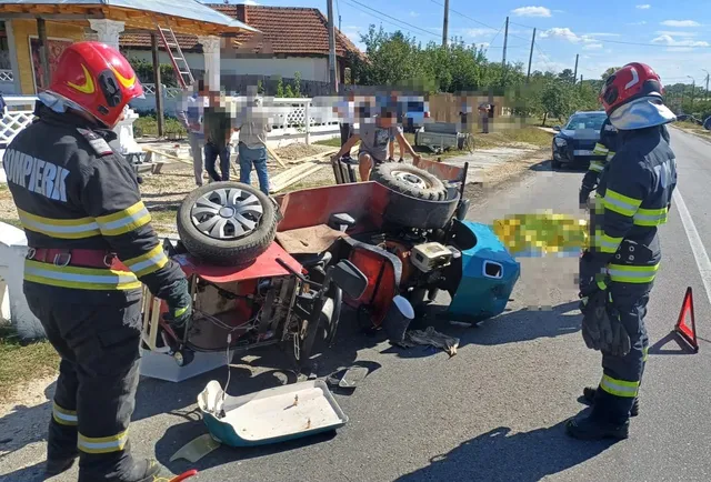 Două persoane în uniforme de pompier stau lângă resturile unui vehicul distrus pe un drum asfaltat.