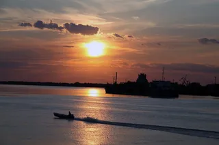 O barcă mică navighează pe un fluviu sub un cer la apus, având siluete de nave și infrastructură portuară în fundal.