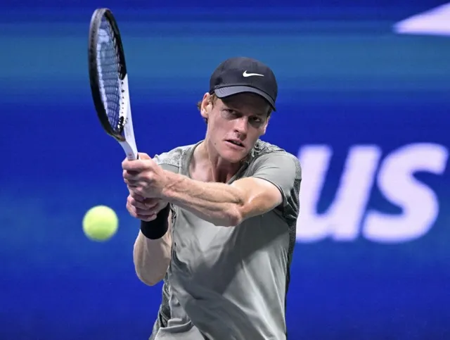Un bărbat cu șapcă și tricou gri lovește o minge de tenis cu o racletă pe un teren albastru.