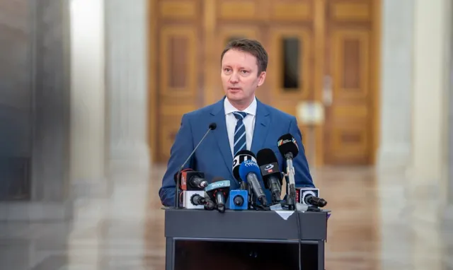 Un bărbat într-un costum albastru și cravată stă în spatele unui podium pe care sunt amplasate mai multe microfoane de presă.