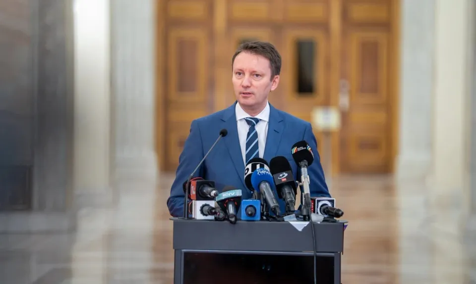 Un bărbat într-un costum albastru și cravată stă în spatele unui podium pe care sunt amplasate mai multe microfoane de presă.