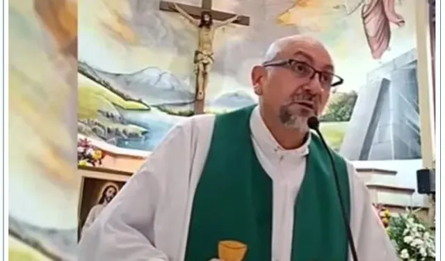 Un bărbat cu ochelari și barbă scurtă poartă o robă albă cu o eșarfă verde, stând în fața unui perete pictat cu peisaje și un crucifix.