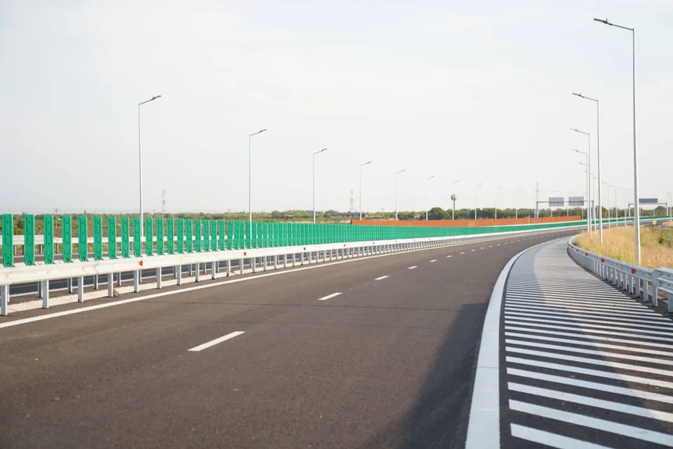 O autostradă cu asfalt negru, linii albe marcate și parapeti metalici este delimitată de un gard verde și stâlpi de iluminat înalți sub un cer deschis.