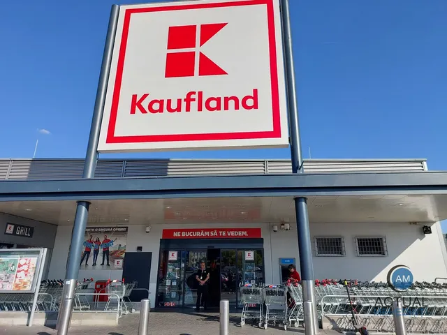 Un semn cu logo-ul Kaufland este montat deasupra intrării unui magazin, unde se văd cărucioare de cumpărături și persoane în fața ușilor vitrate.
