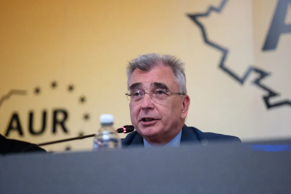 Un bărbat cu ochelari și părul cărunt stă la o masă în fața unui fundal cu literele AUR.