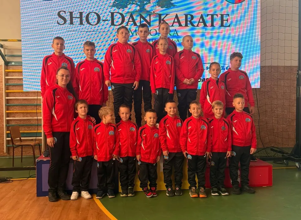 Un grup de copii și adulți îmbrăcați în treninguri roșii stau în rând în fața unui ecran mare cu textul SHO-DAN KARATE.