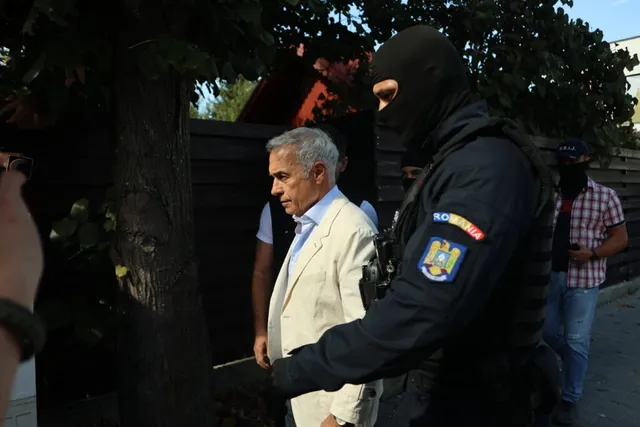 Un bărbat cu părul cărunt și sacou deschis este însoțit de un agent în uniformă neagră și mască.