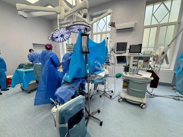 Personal medical în halate albastre efectuează o procedură într-o sală de operație dotată cu echipamente tehnice și lumini chirurgicale suspendate.