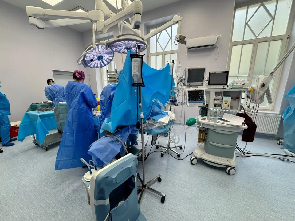 Personal medical în halate albastre efectuează o procedură într-o sală de operație dotată cu echipamente tehnice și lumini chirurgicale suspendate.