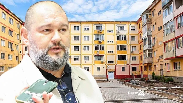 Un bărbat cu barbă și ochelari de soare agâțați de cămașă ține un telefon mobil în fața unor blocuri de apartamente galbene și portocalii.