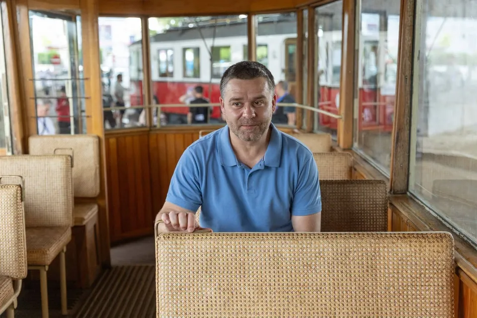Un bărbat cu tricou polo albastru stă în interiorul unui vehicul cu ferestre mari și scaune cu spătar textil bej.