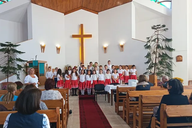 Un grup de copii în uniformă albă și roșie stă în rând în fața unui cadru didactic într-o încăpere cu un cruce pe perete.