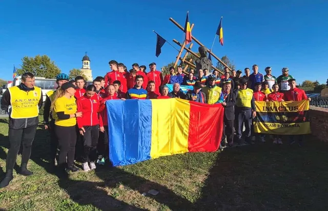 Un grup de persoane în picioare pe un ierpet, ținând steagul României și un steag galben, înaintea unui fundal cu un turn și o construcție de lemn.