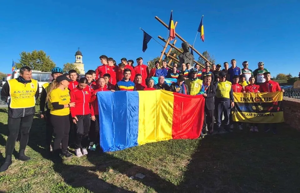 Un grup de persoane în picioare pe un ierpet, ținând steagul României și un steag galben, înaintea unui fundal cu un turn și o construcție de lemn.