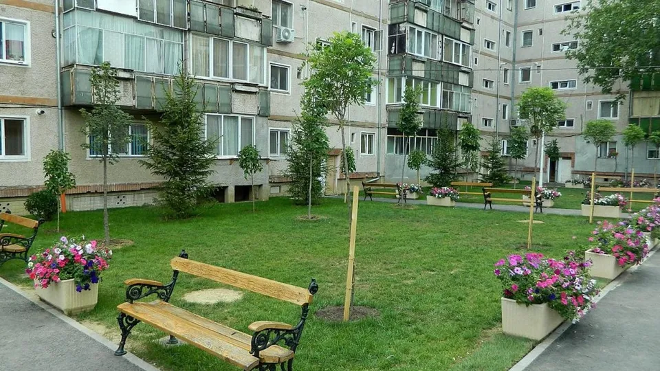 Un spațiu verde cu iarbă, bănci de lemn și ghivece cu flori colorate este situat în fața unor clădiri de locuințe înalte.