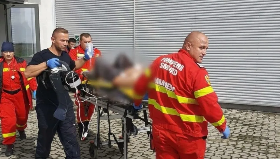 Patru persoane transportă un brancard pe care se află o persoană, în timp ce doi bărbați poartă uniforme roșii cu benzi galbene reflectorizante.