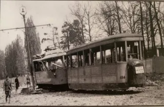 Două tramvaie avariate stau pe șine lângă un stâlp de iluminat, în fața unor copaci fără frunze.
