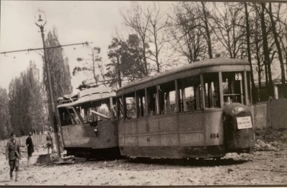 Două tramvaie avariate stau pe șine lângă un stâlp de iluminat, în fața unor copaci fără frunze.