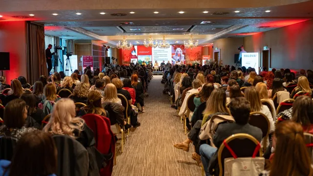 O mulțime de persoane stă așezată pe scaune într-o sală de conferințe, orientată spre o scenă cu un ecran mare și un podium iluminat.